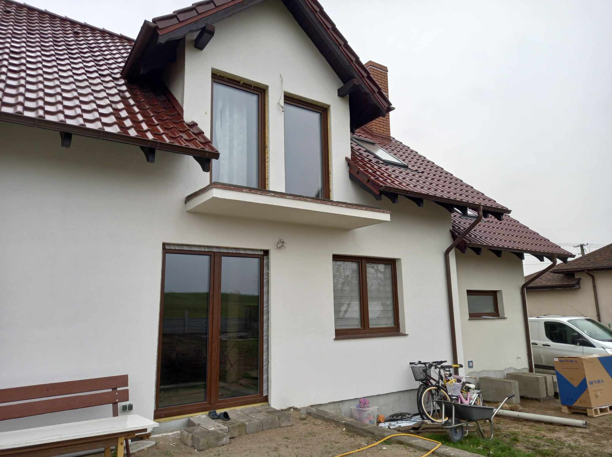 Biały dom jednorodzinny z brązowymi ramami okiennymi i drzwiami przesuwnymi, widoczne elementy montażowe i rower zaparkowany obok ściany.