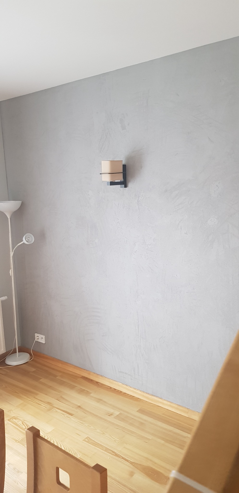 Ściana pomalowana farbą dekoracyjną w odcieniach szarości z zamontowanym kinkietem, widoczna lampa podłogowa i fragment drewnianej podłogi oraz krzesła.