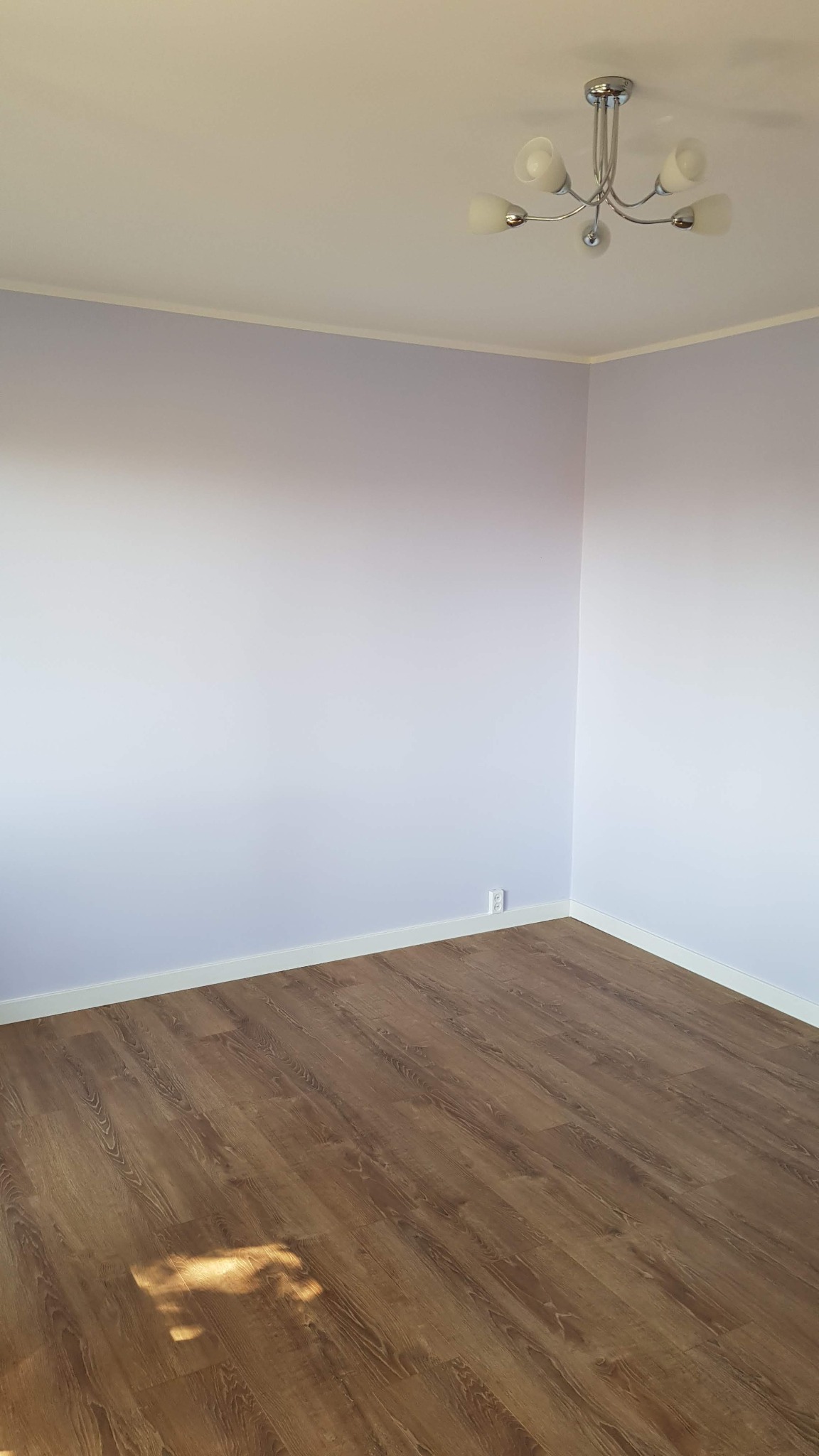 Pomalowane na fioletowo ściany w pustym pokoju z ułożonymi panelami podłogowymi, widoczny nowoczesny żyrandol sufitowy.