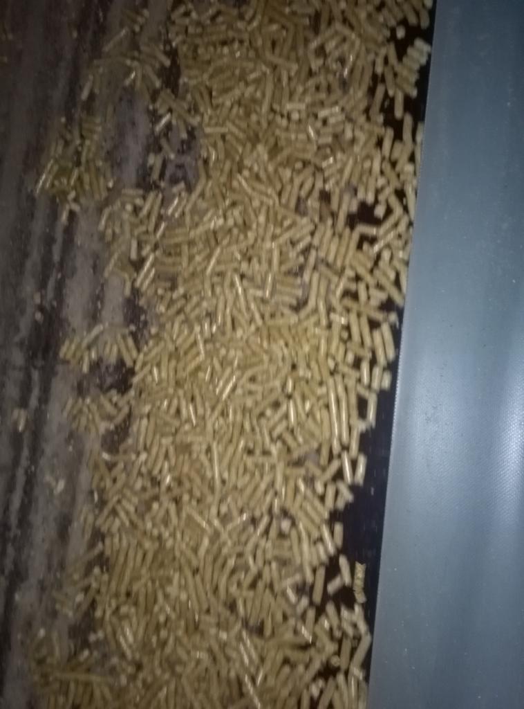 Jasne, drewniane pellety rozsypane na ciemnej powierzchni, widoczne drobne zabrudzenia i fragment przezroczystej folii w prawym dolnym rogu.