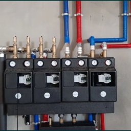 INSTAL GrOs - Rozdzielacz ogrzewania podłogowego z czterema obiegami, widoczne manometry, elektroniczne sterowniki, rury w kolorach czerwonym i niebieskim, izolowane rury pionowe.