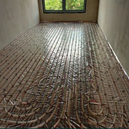 INSTAL GrOs - Rozłożona instalacja ogrzewania podłogowego Reflex Thermosystem na folii izolacyjnej z czerwoną siatką, widok z góry, surowy stan wykończenia pomieszczenia.