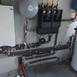 Kotłownia z piecem gazowym Termet, zbiornikami wyrównawczymi, rozdzielaczem Oventrop i rurami grzewczymi z izolacją, widoczne manometry i zawory.