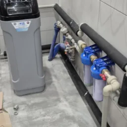 Szary zmiękczacz wody obok instalacji z dwoma filtrami mechanicznymi, rurami w izolacji termicznej i zaworami, całość na płytkach imitujących beton w pomieszczeniu z białą glazurą.