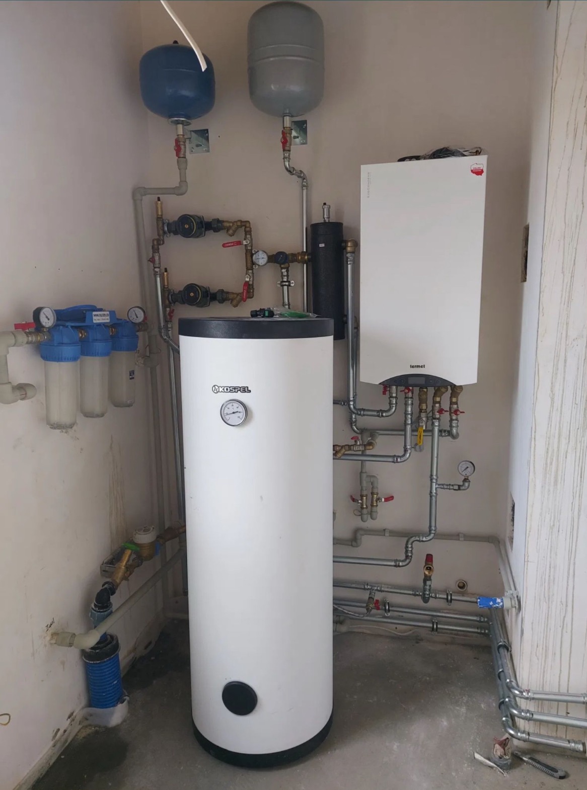 Zestaw instalacji grzewczej z kotłem Termet, zasobnikiem Kospel, zbiornikami wyrównawczymi oraz filtrami wody, zamontowany w kotłowni z surowymi ścianami.