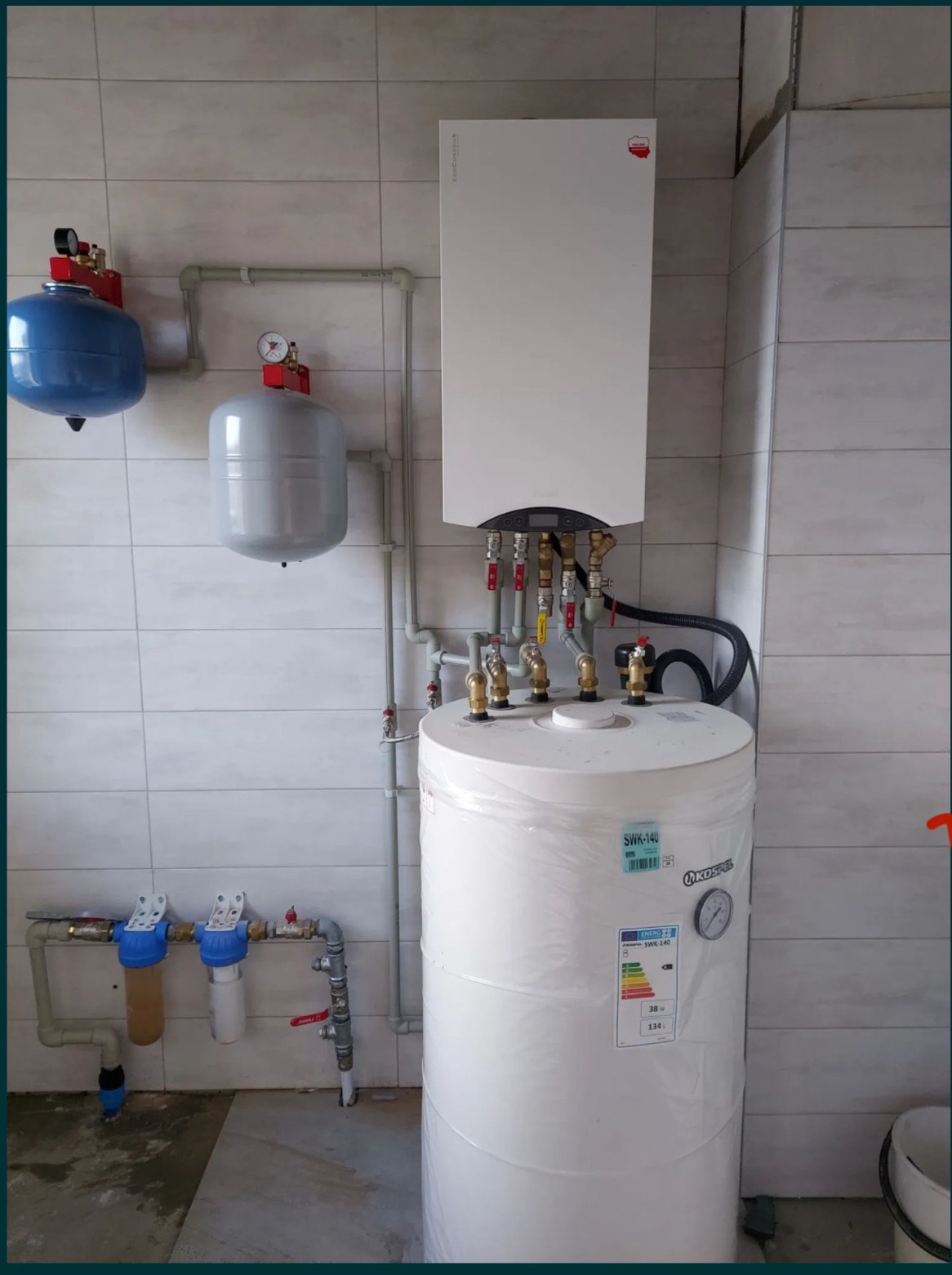 Biały, wiszący piec gazowy marki Kospel, podłączony do pionowego, owiniętego folią ochronną zasobnika ciepłej wody, z widocznymi manometrami, zaworami i filtrami wody na tle jasnych, kwadratowych...