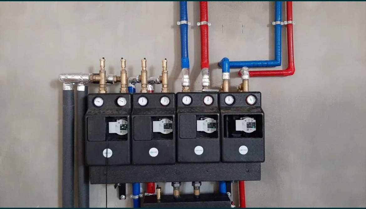 Rozdzielacz ogrzewania podłogowego z czterema obiegami, widoczne manometry, elektroniczne sterowniki, rury w kolorach czerwonym i niebieskim, izolowane rury pionowe.