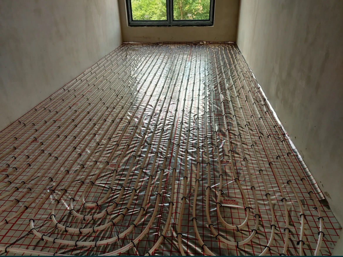 Rozłożona instalacja ogrzewania podłogowego Reflex Thermosystem na folii izolacyjnej z czerwoną siatką, widok z góry, surowy stan wykończenia pomieszczenia.