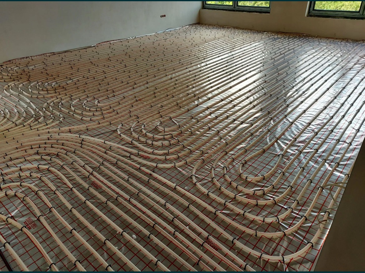 Rozłożona instalacja ogrzewania podłogowego na folii z czerwoną siatką, widoczne rury w kształcie meandrów, jasne wnętrze z oknami w tle.