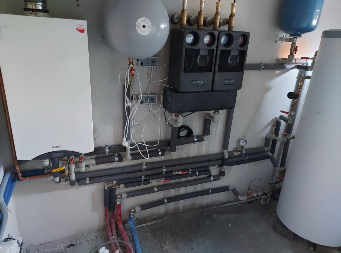 Kotłownia z piecem gazowym Termet, zbiornikami wyrównawczymi, rozdzielaczem Oventrop i rurami grzewczymi z izolacją, widoczne manometry i zawory.