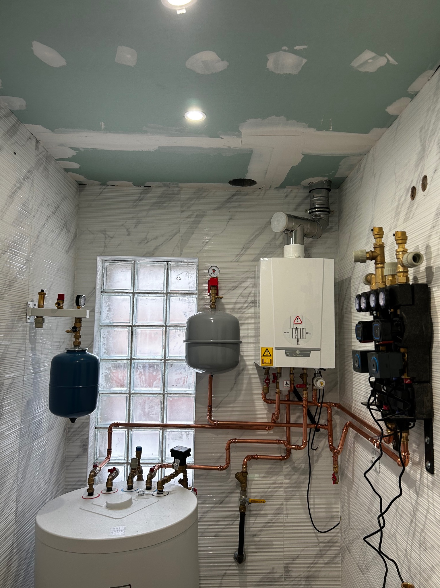 Kotłownia w trakcie montażu: biały piec gazowy, zbiornik akumulacyjny, naczynia przeponowe, rozdzielacz, rury miedziane, ściany wykończone płytkami imitującymi marmur, sufit w trakcie wykańczania...