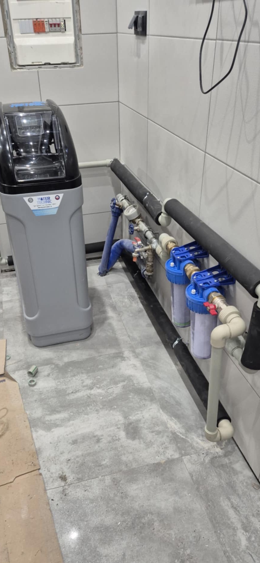 Szary zmiękczacz wody obok instalacji z dwoma filtrami mechanicznymi, rurami w izolacji termicznej i zaworami, całość na płytkach imitujących beton w pomieszczeniu z białą glazurą.