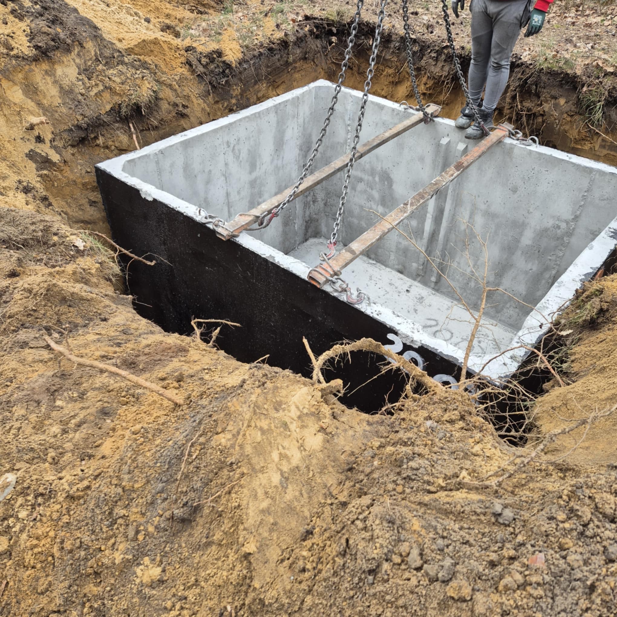 Betonowy zbiornik opuszczany do wykopu za pomocą łańcuchów, widoczne drewniane belki stabilizujące, ziemia wokół wykopu, pracownik w tle.