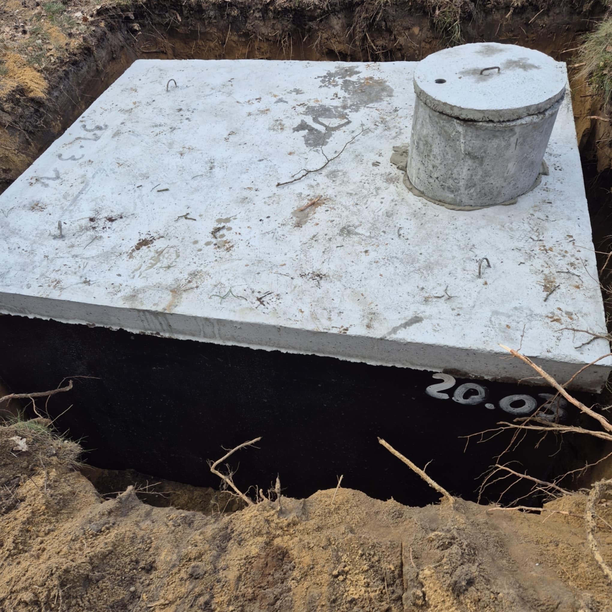Betonowy zbiornik, prawdopodobnie szambo, umieszczony w wykopie, z widoczną pokrywą włazu i czarną izolacją. Na izolacji widnieją białe cyfry 20.0.