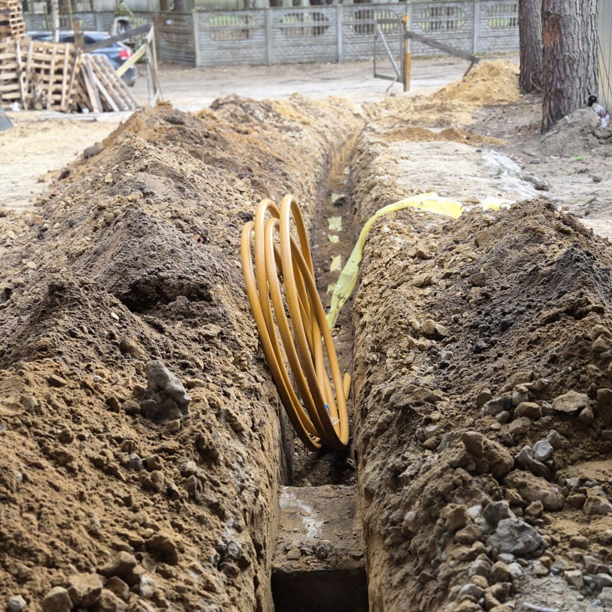 Wykop w ziemi z żółtym przewodem wewnątrz, widoczna żółta taśma ostrzegawcza, w tle ogrodzenie posesji i palety.