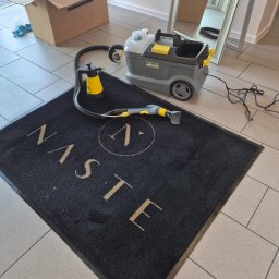PURO Częstochowa - Czarny dywanik z beżowym napisem 'NASTE' i logo, obok odkurzacz piorący Karcher, karton i lustro. Sprzęt do czyszczenia dywanów w pomieszczeniu.