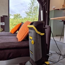 PURO Częstochowa - Profesjonalny odkurzacz piorący Kärcher na tle nowoczesnego salonu z dużym oknem i brązową kanapą z pomarańczowymi poduszkami. Widoczne ślady po czyszczeniu.