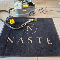 PURO Częstochowa - Czarny dywanik z napisem 'NASTE' czyszczony odkurzaczem piorącym Karcher na jasnych płytkach podłogowych. Widoczny fragment osoby w prawym górnym rogu.