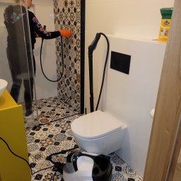 PURO Częstochowa - Osoba czyści parą kabinę prysznicową w łazience z wzorzystymi płytkami, używając parownicy stojącej obok toalety. Widoczny także środek czyszczący.