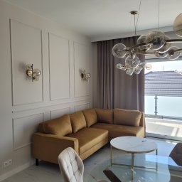 PURO Częstochowa - Stylowy salon z narożną sofą w odcieniach złota, marmurową podłogą i designerskim oświetleniem. Widok na balkon przez okno z szarymi zasłonami.