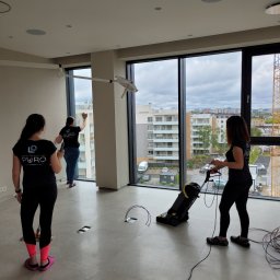 PURO Częstochowa - Ekipa sprzątająca Puro w akcji: mycie okien i odkurzanie podłogi w przestronnym, nowoczesnym wnętrzu z widokiem na miasto. Widoczny sprzęt i logo firmy.