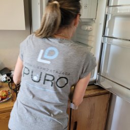 PURO Częstochowa - Osoba w koszulce firmy sprzątającej czyści wnętrze lodówki, otwierając drzwi. Widoczna zawartość lodówki i drewniana szafka. Na blacie stoi talerz z jedzeniem.