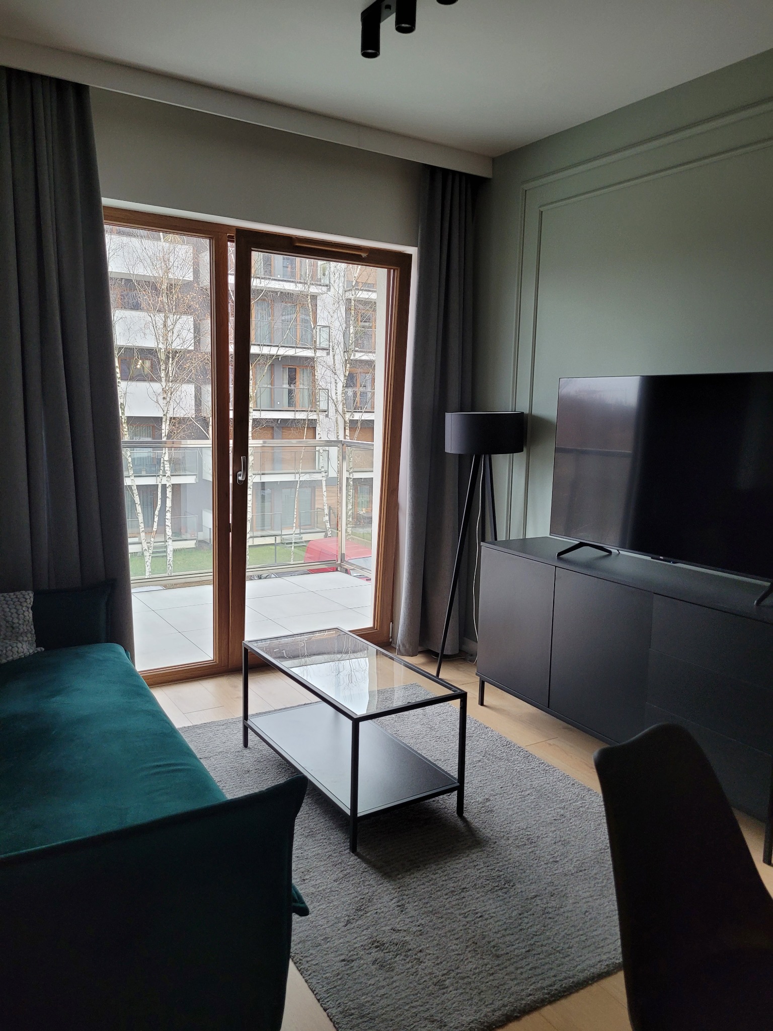 Nowoczesny salon z balkonem: szara sofa, szklany stolik, TV na szafce. Widok na osiedle z drzewami przez duże okno balkonowe.