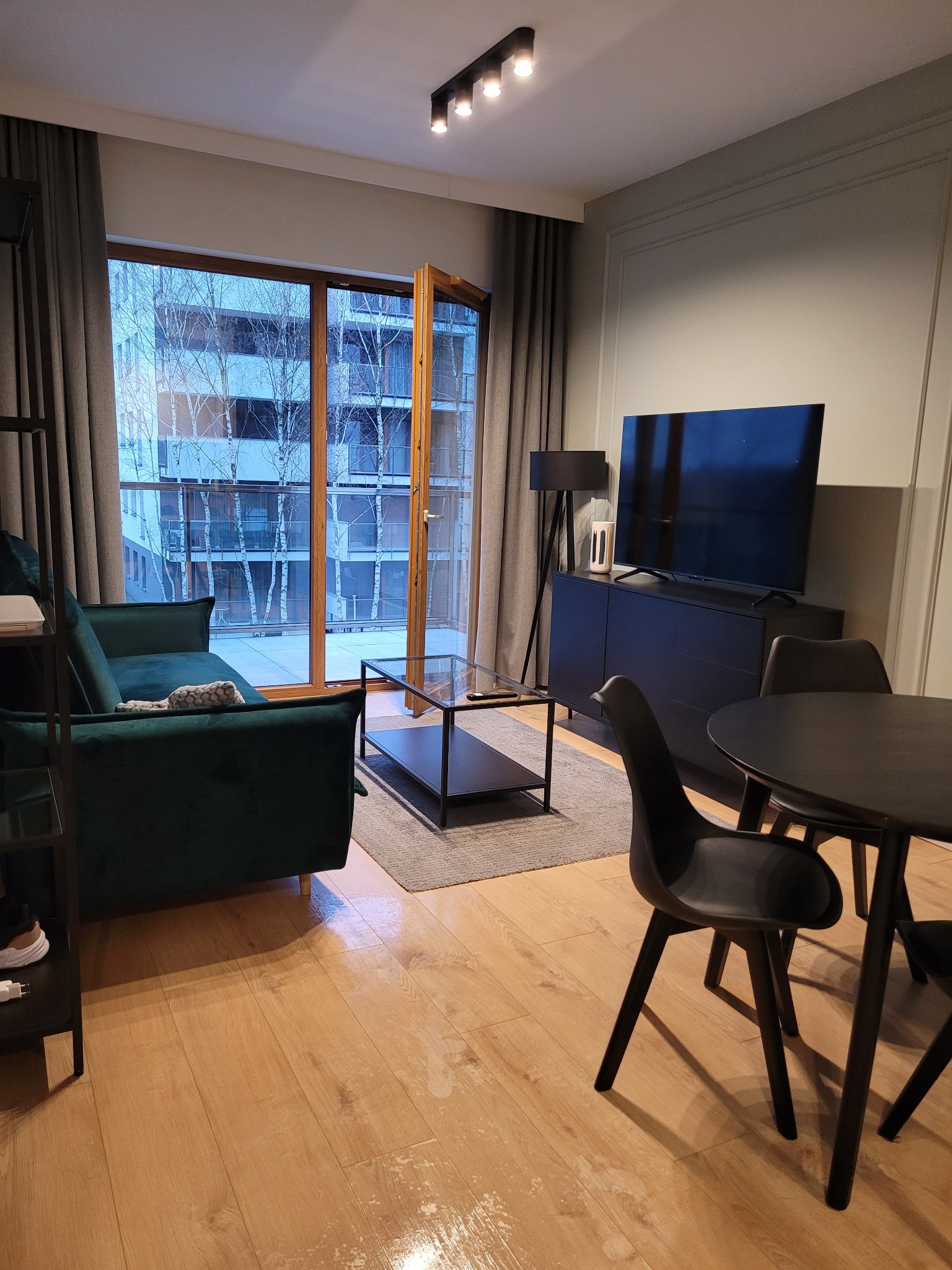 Nowoczesny salon z zieloną sofą, czarnym stolikiem, stołem i krzesłami na tle okna balkonowego z widokiem na budynek. Parkiet z połyskiem.