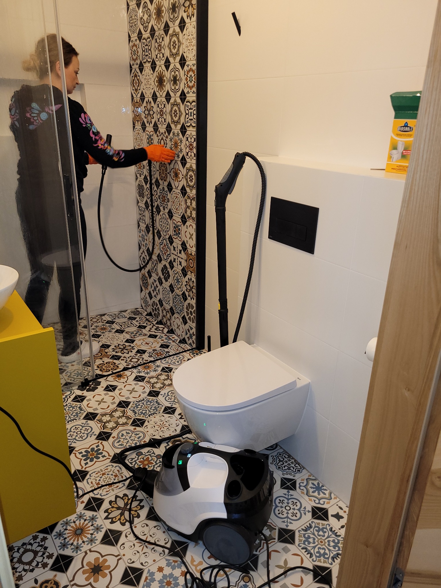 Osoba czyści parą kabinę prysznicową w łazience z wzorzystymi płytkami, używając parownicy stojącej obok toalety. Widoczny także środek czyszczący.