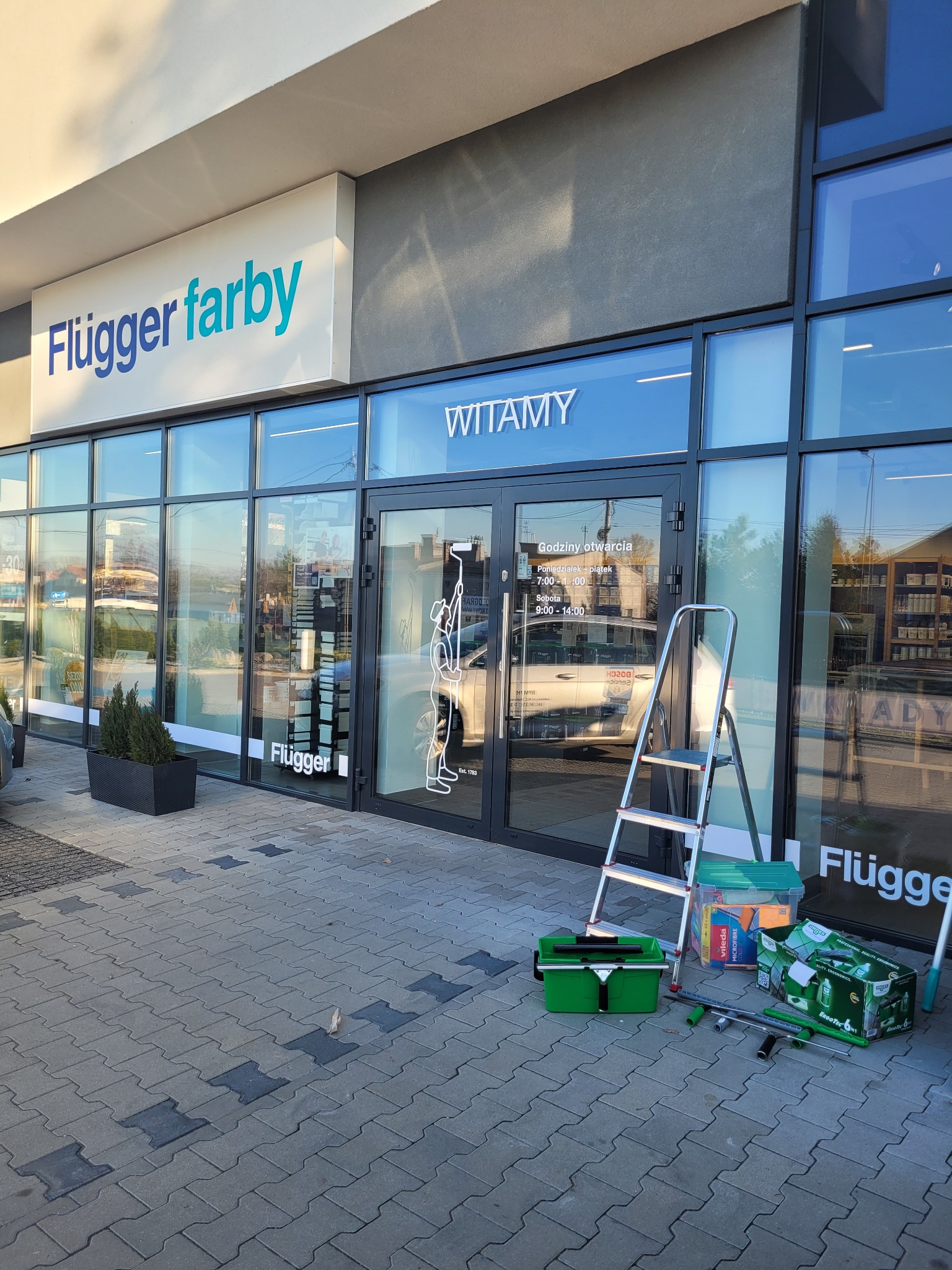 Mycie okien w Częstochowie. Drabina i akcesoria do czyszczenia szyb przed witryną sklepu z farbami Flügger. Widoczne godziny otwarcia na szybie.