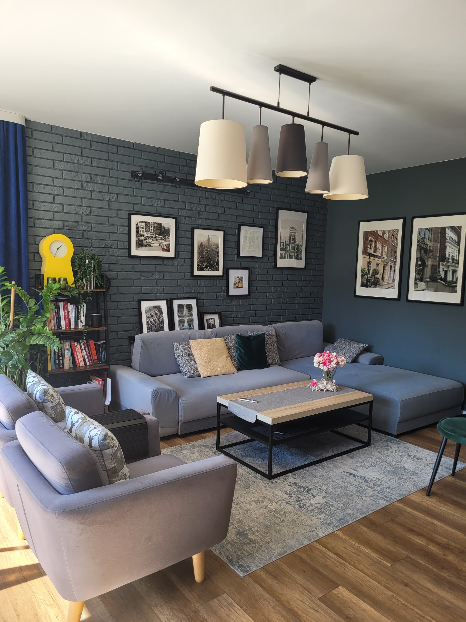 Stylowy salon z szarą sofą narożną, ceglaną ścianą i dekoracyjnymi obrazami. Na stoliku kwiaty, a obok regał z książkami i żółtym zegarem. Przytulne wnętrze.