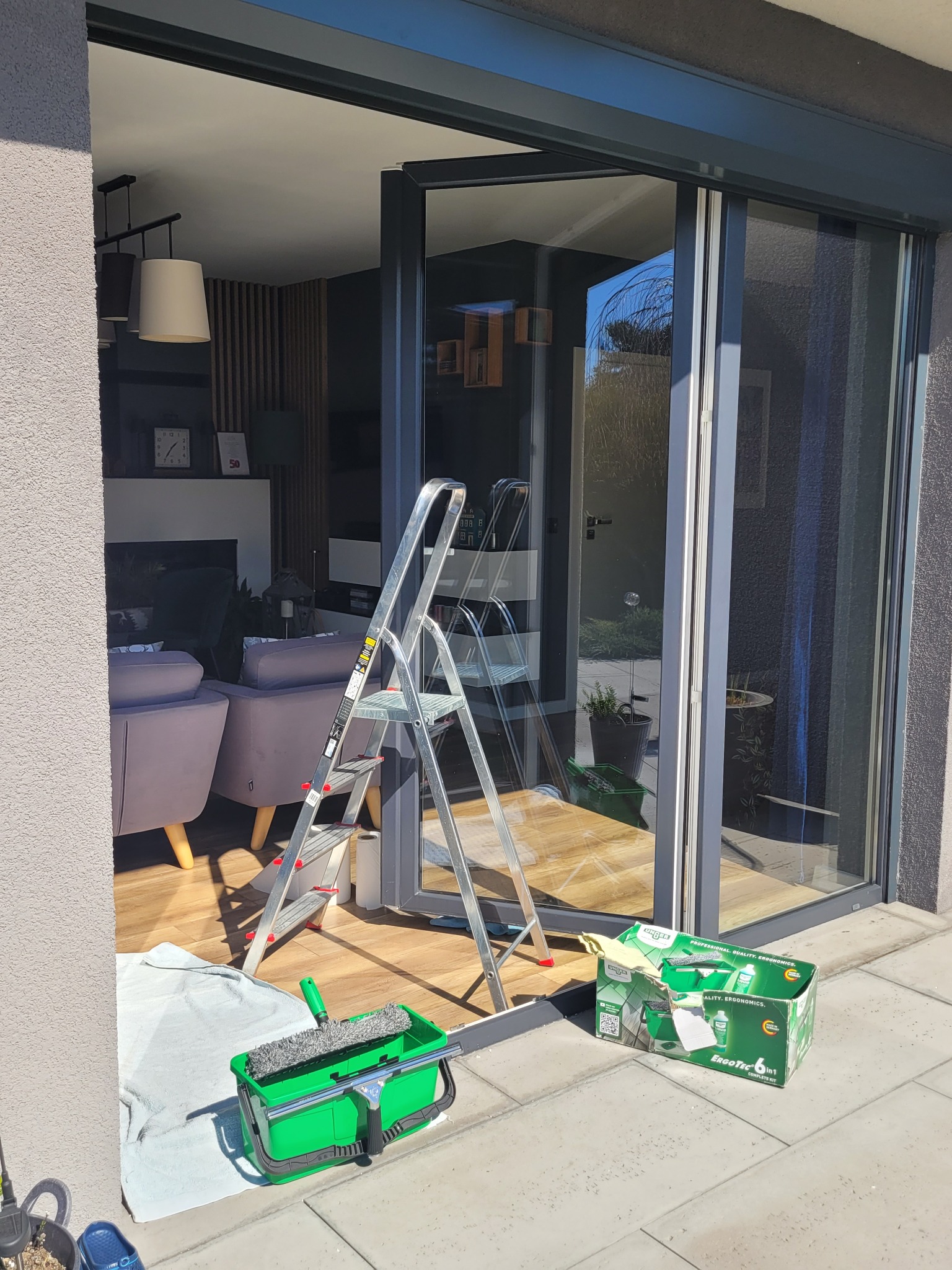 Aluminiowa drabina oparta o przesuwne szklane drzwi tarasowe. Obok wiadro z myjką i karton z zestawem do mycia okien. W tle widoczny salon z szarymi fotelami.