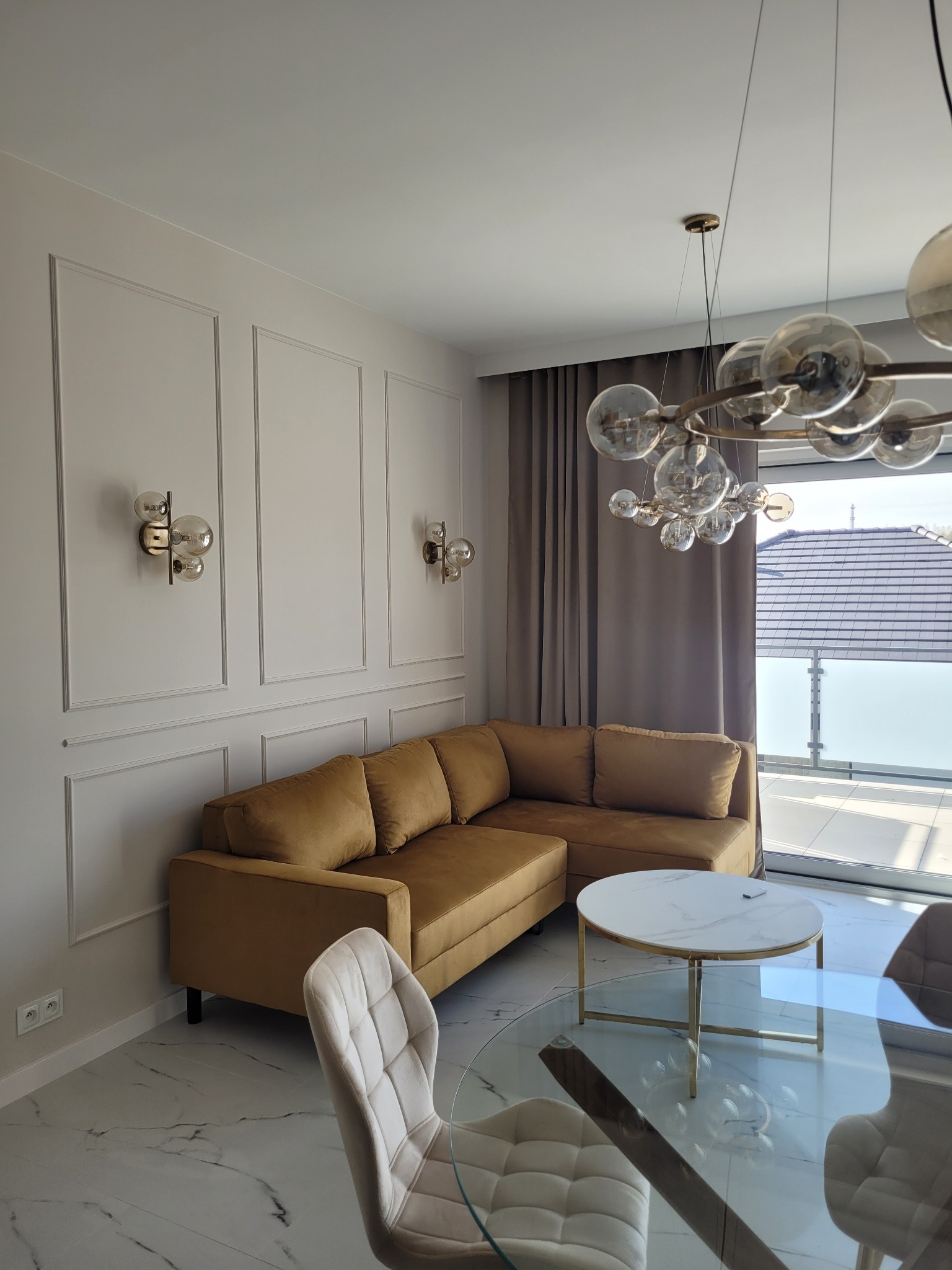 Stylowy salon z narożną sofą w odcieniach złota, marmurową podłogą i designerskim oświetleniem. Widok na balkon przez okno z szarymi zasłonami.