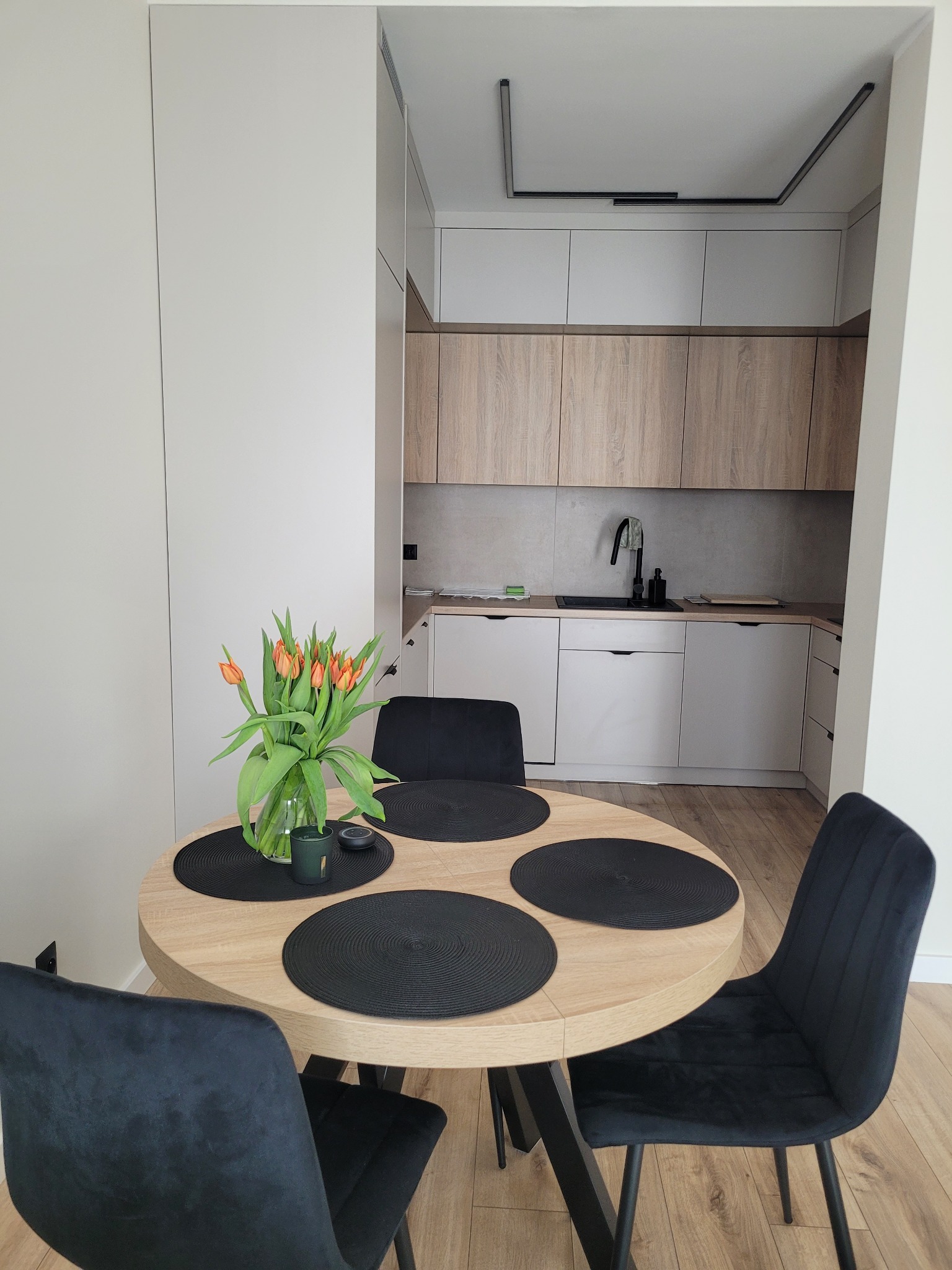 Jasna kuchnia z drewnianymi szafkami i okrągłym stołem z czarnymi podkładkami. Na stole wazon z pomarańczowymi tulipanami i czarne krzesła.