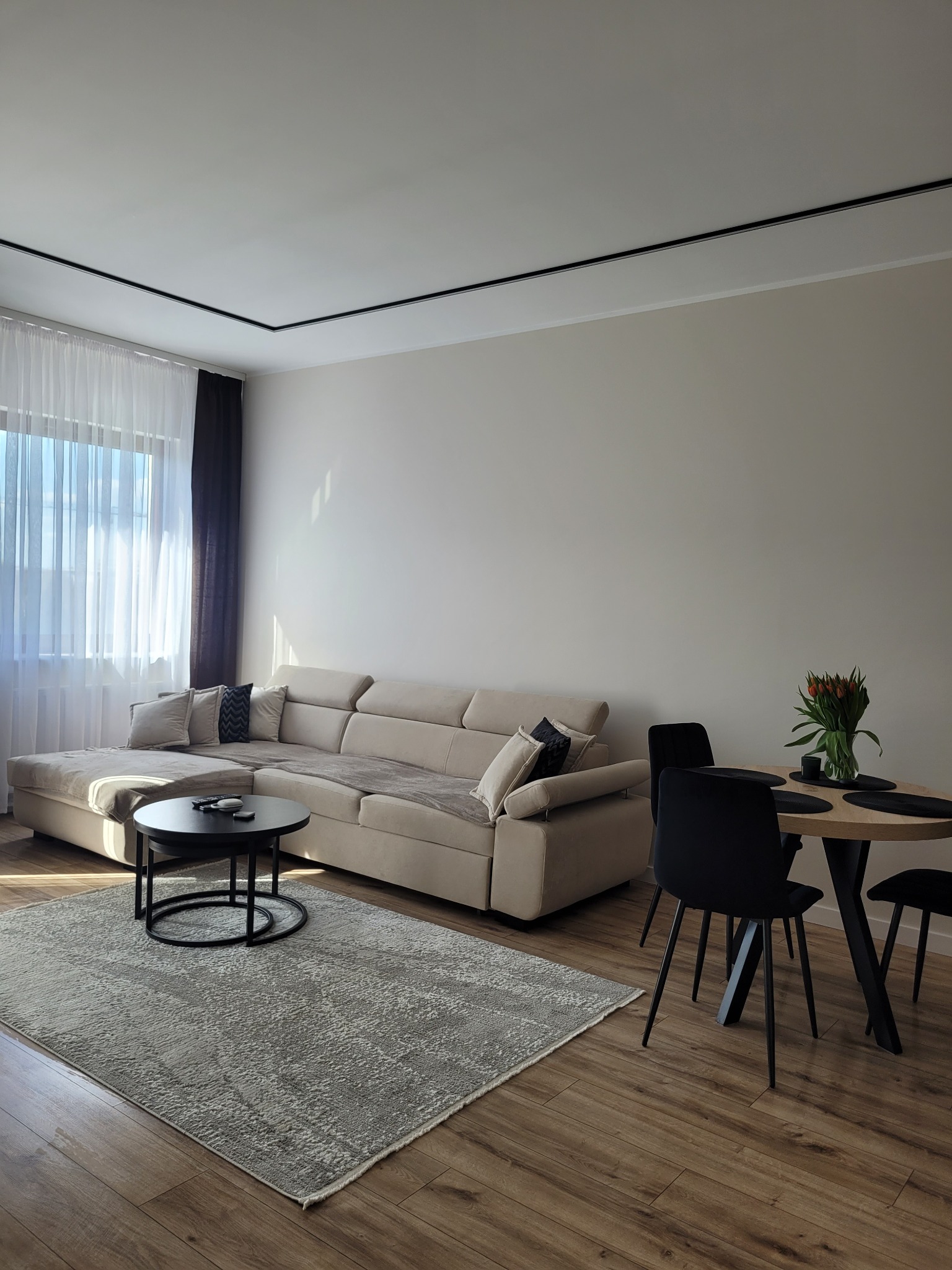 Jasny salon z beżową sofą, okrągłym stolikiem kawowym, dywanem i stołem z czarnymi krzesłami. W wazonie tulipany, światło słoneczne wpada przez okno.