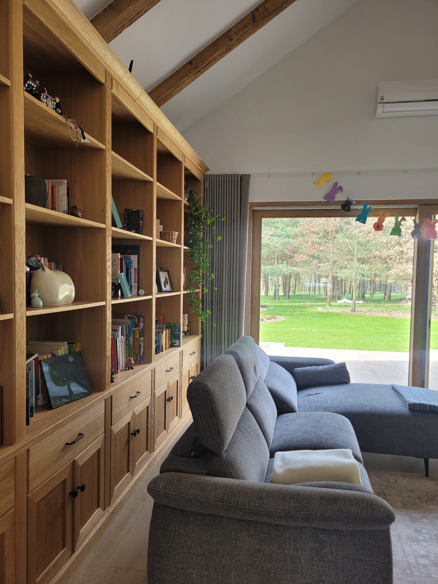 Jasny salon z dębowym regałem zabudowanym, szarą kanapą i widokiem na ogród przez okno. Dekoracje wielkanocne wiszą przy oknie, dodając wnętrzu wiosenny akcent.