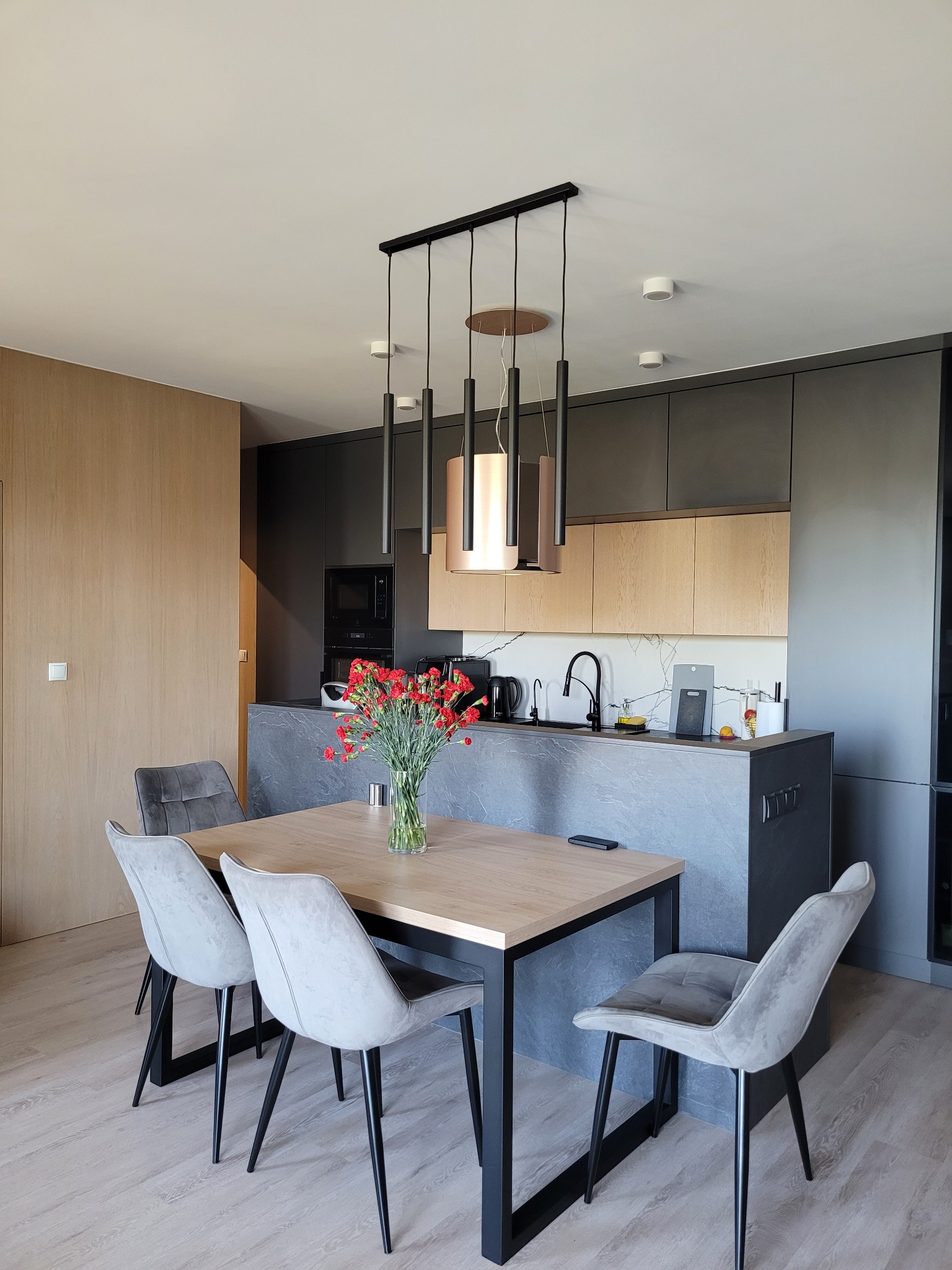 Nowoczesna kuchnia z jadalnią: szare szafki, drewniane akcenty, marmurowy blat, stół z kwiatami i designerskie lampy wiszące. Minimalistyczny, ale przytulny wystrój.