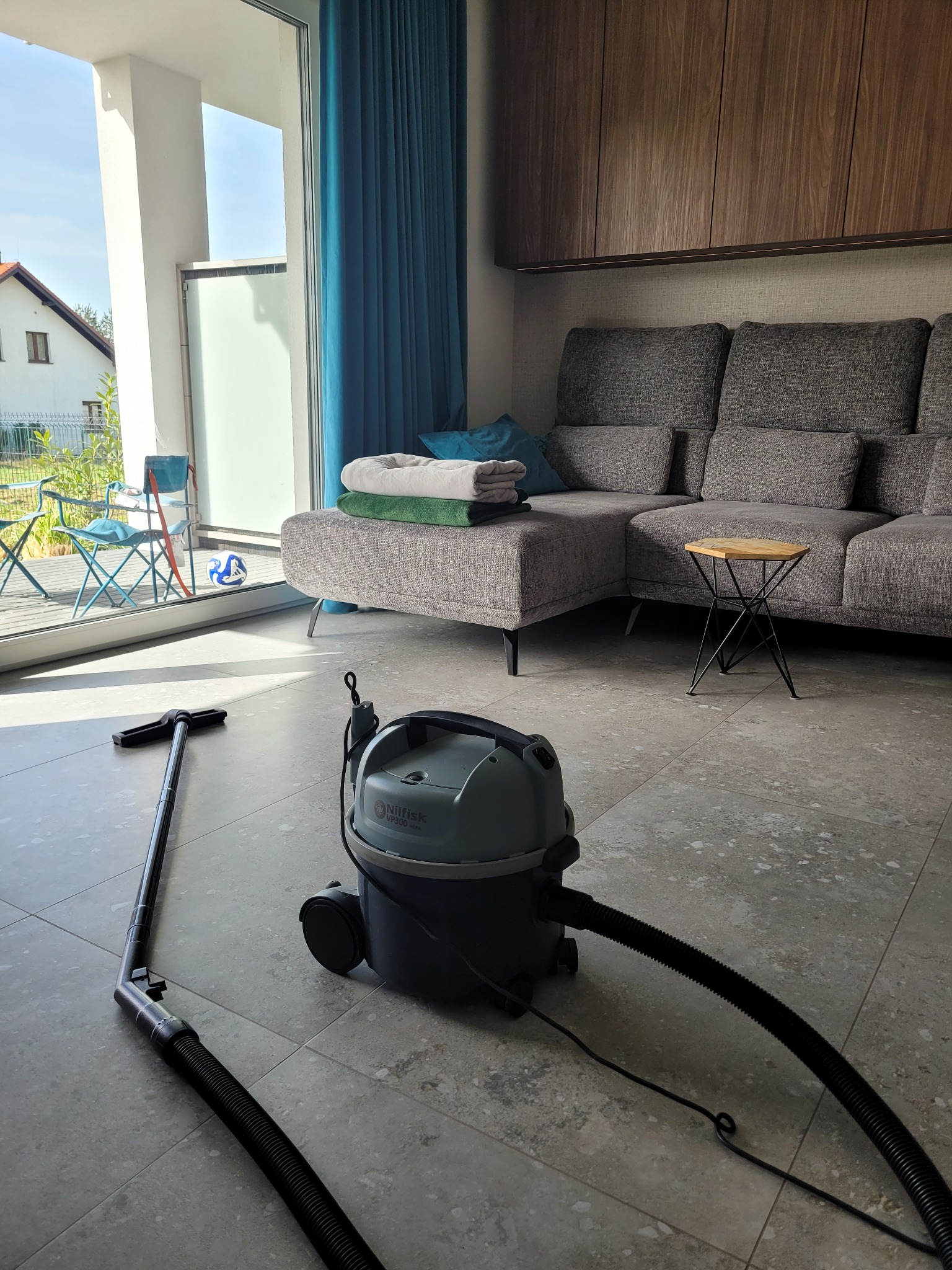 Szary odkurzacz Nilfisk VP300 na kafelkach w salonie. W tle sofa, stolik, balkon z widokiem na dom i niebieskie zasłony. Sprzęt do sprzątania w nowoczesnym wnętrzu.