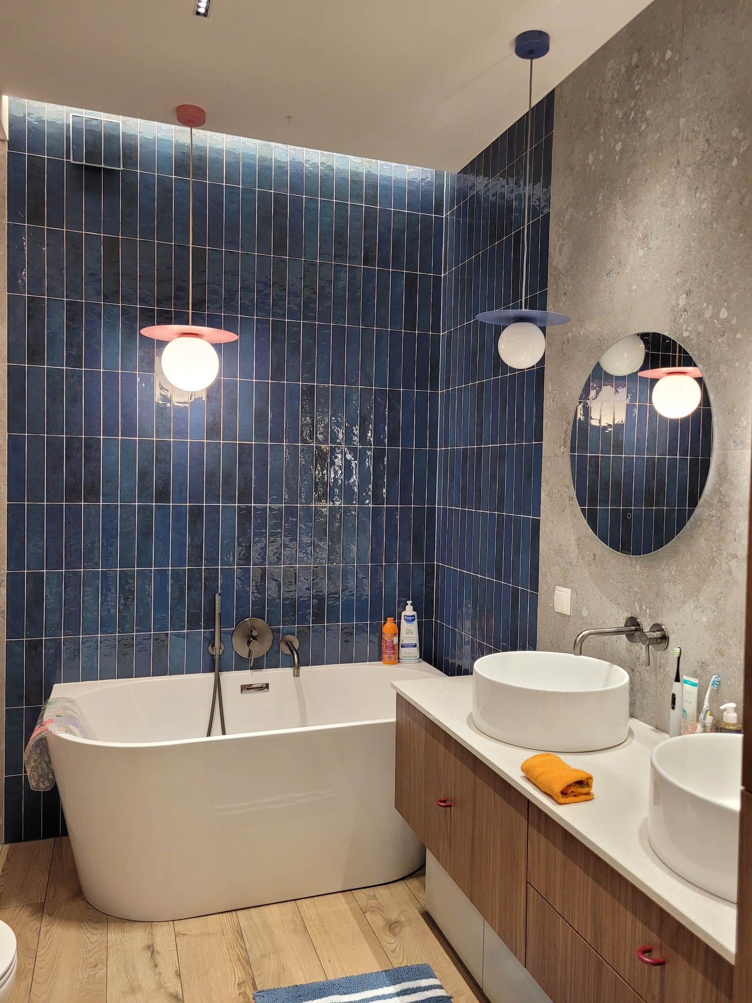 Nowoczesna łazienka z wanną, umywalkami nablatowymi i niebieskimi płytkami. Dwie lampy wiszące z kolorowymi akcentami. Drewnopodobna podłoga i szafka pod umywalką.