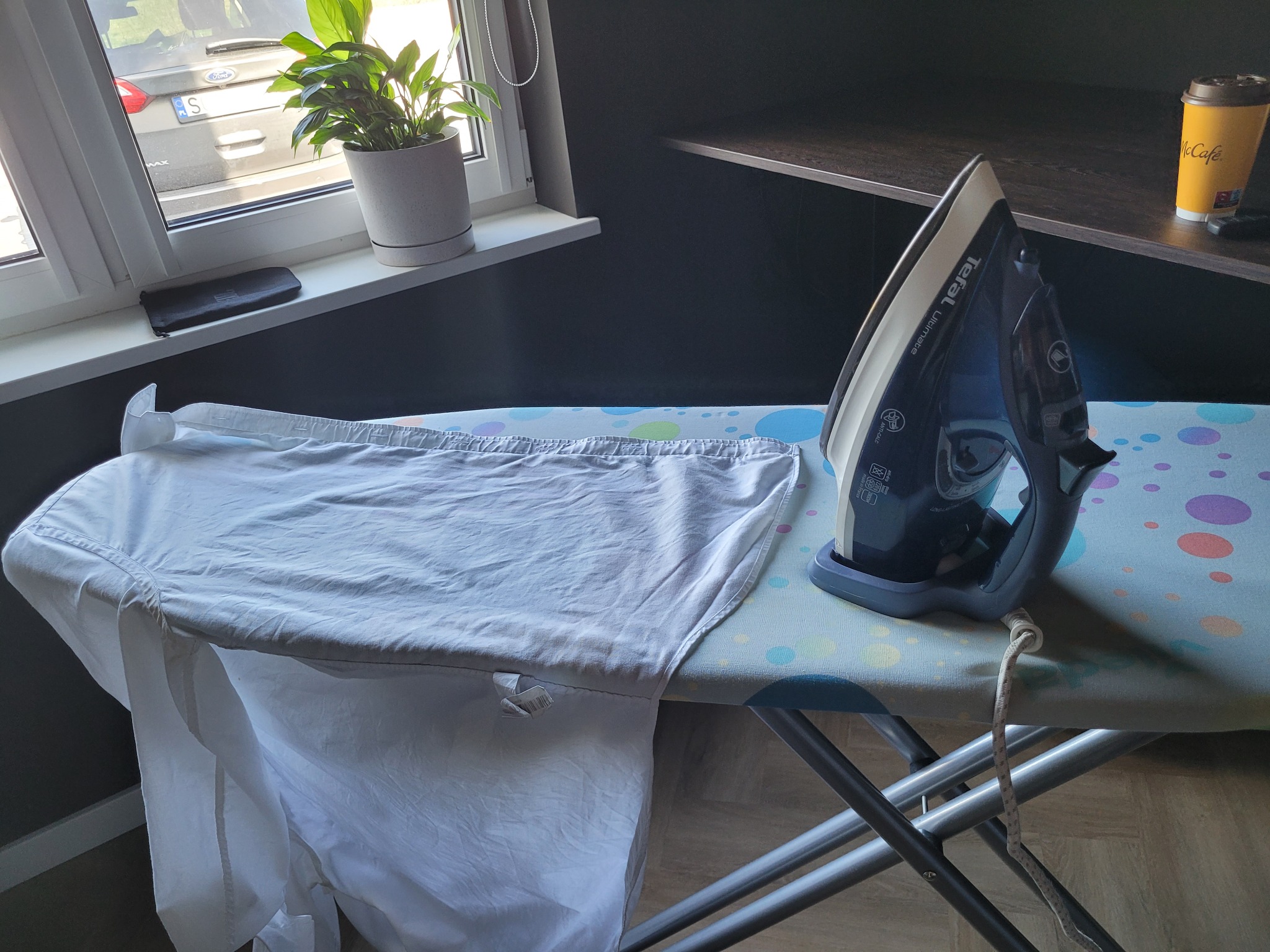 Żelazko Tefal na desce do prasowania z koszulą. W tle okno, roślina w doniczce i półka. Scena domowa, wnętrze.