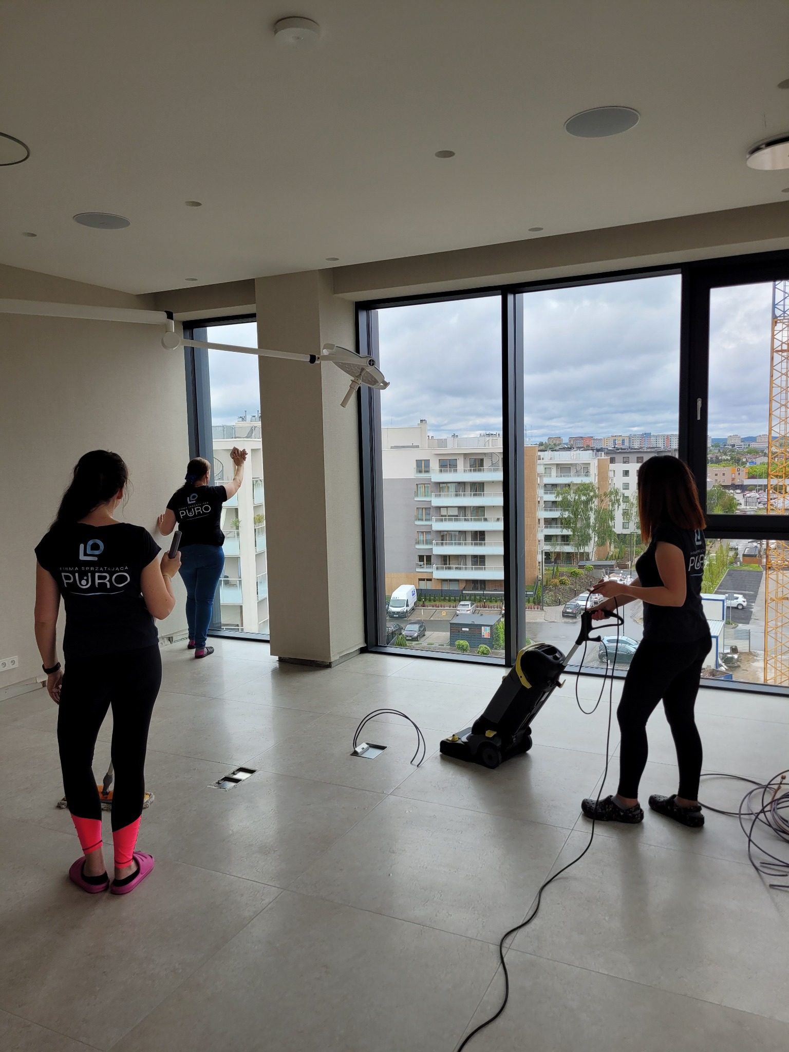 Ekipa sprzątająca Puro w akcji: mycie okien i odkurzanie podłogi w przestronnym, nowoczesnym wnętrzu z widokiem na miasto. Widoczny sprzęt i logo firmy.