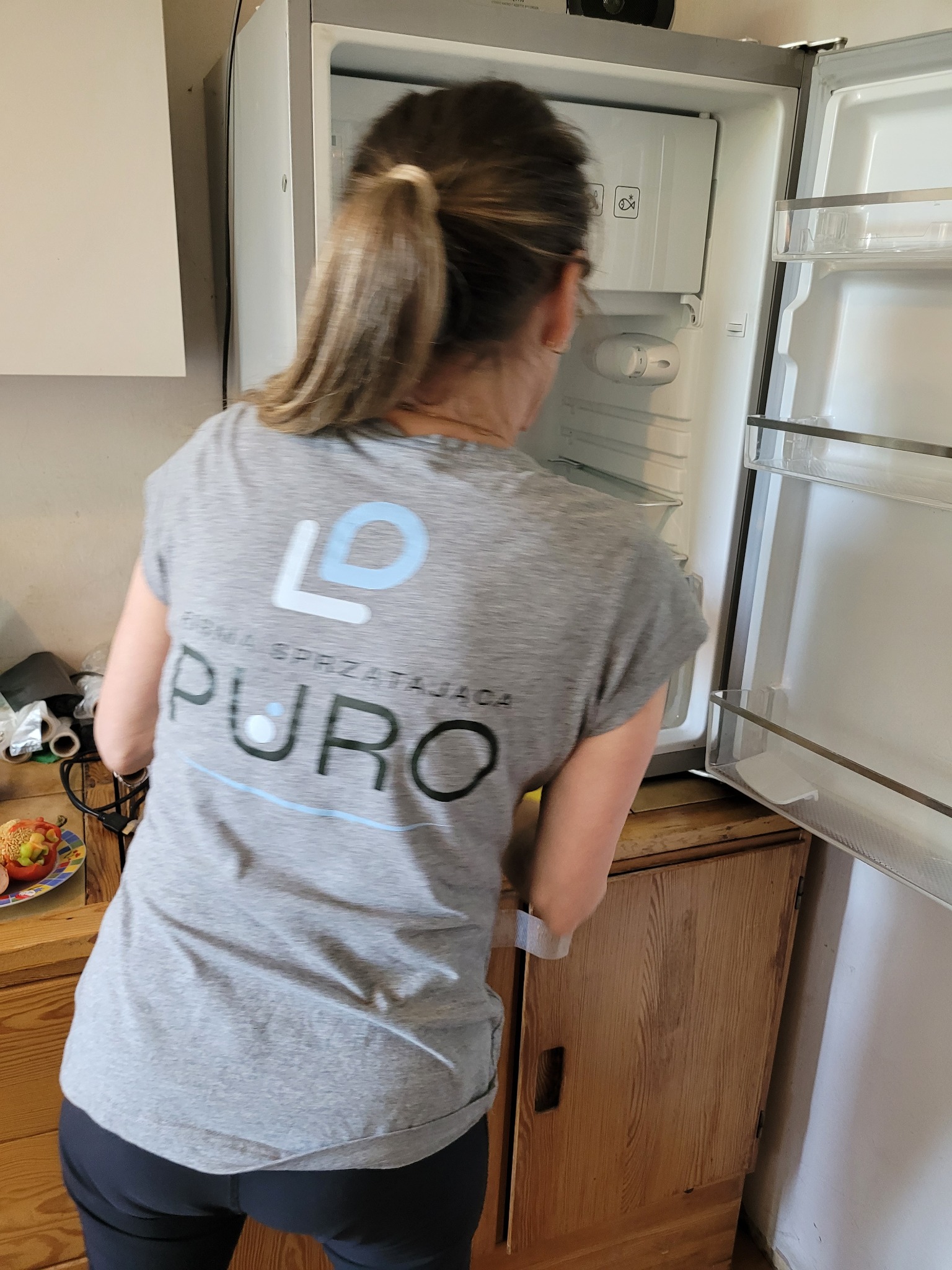 Osoba w koszulce firmy sprzątającej czyści wnętrze lodówki, otwierając drzwi. Widoczna zawartość lodówki i drewniana szafka. Na blacie stoi talerz z jedzeniem.