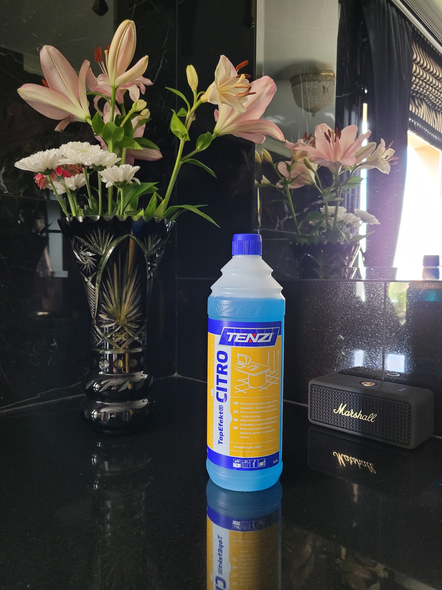 Buteleczka płynu czyszczącego Tenzi Citra stoi na błyszczącym blacie obok wazonu z kwiatami i głośnika Marshall, na tle czarnej ściany z połyskiem.