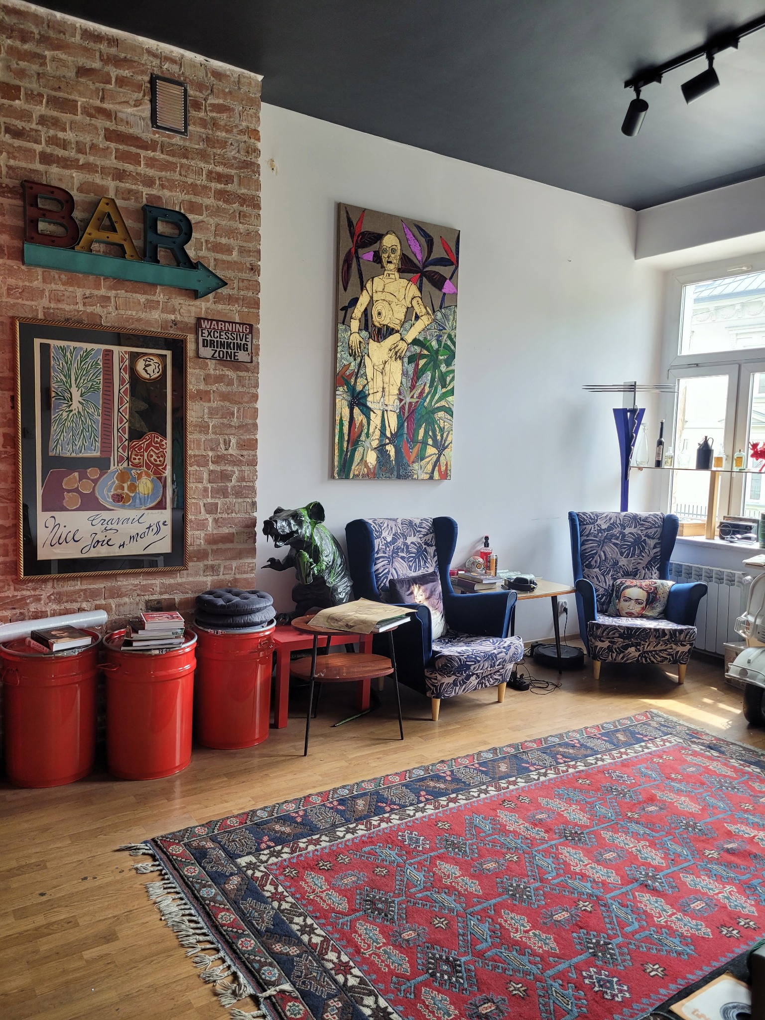 Salon w stylu eklektycznym z ceglaną ścianą, czerwonymi pojemnikami, wzorzystymi fotelami i dywanem orientalnym. Na ścianie grafika i neon 'BAR'.