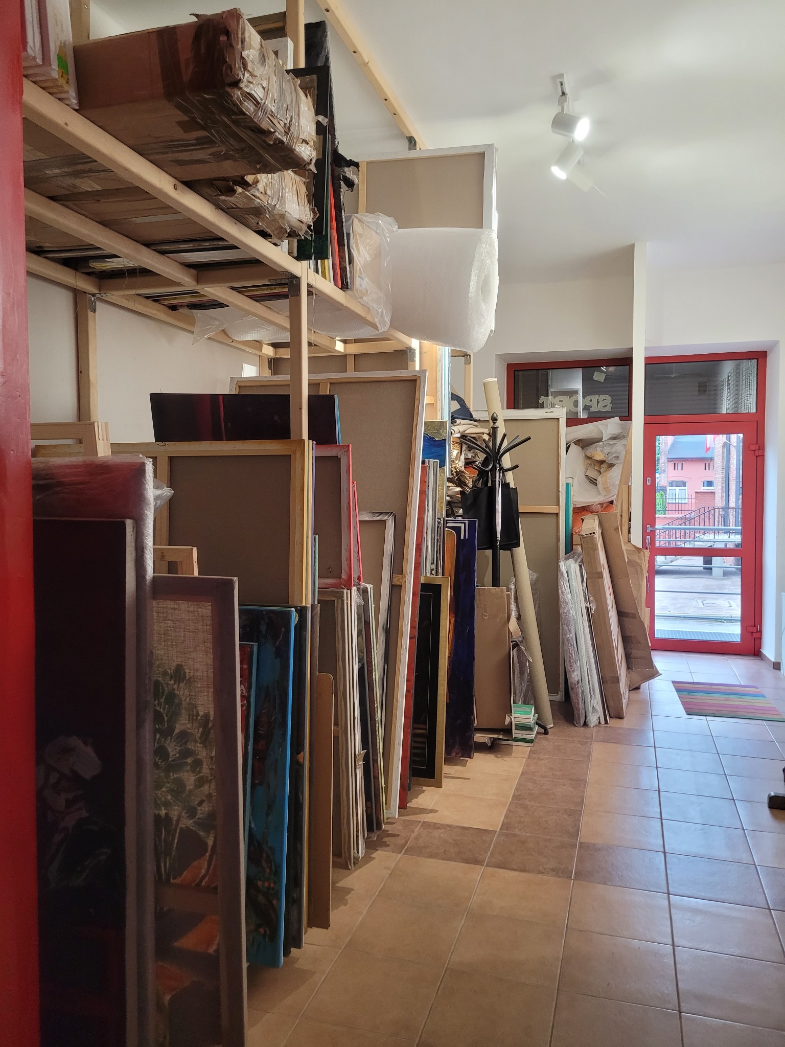 Przechowalnia obrazów w pracowni artystycznej. Ustawione pionowo płótna, niektóre zapakowane, z widocznymi fragmentami malowideł, regał z kartonami i akcesoriami.