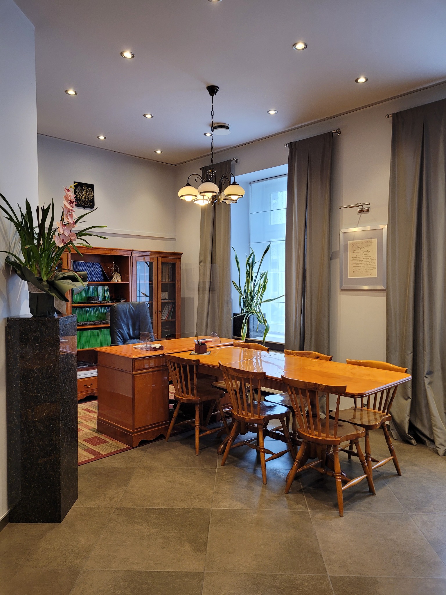 Klimatyczne, jasne biuro z drewnianymi meblami: biurko, krzesła i regał z książkami. Na biurku wazon z kwiatami, w oknie roślina. Szare zasłony i stonowana kolorystyka.