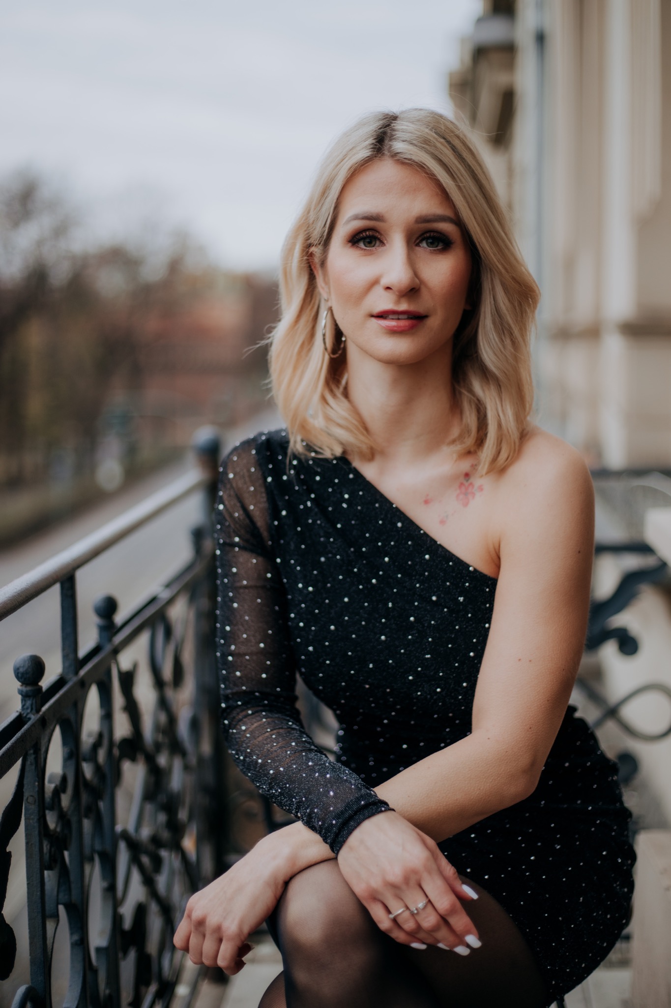 Portret kobiety w czarnej, cekinowej sukience na balkonie z metalową balustradą, z tatuażem kwiatowym na ramieniu i pierścionkami na palcach.