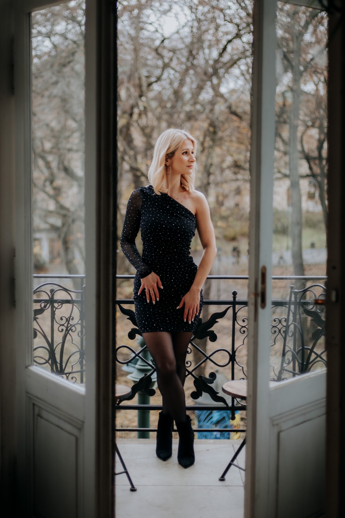 Kobieta w czarnej, cekinowej sukience na jednym ramiączku, stojąca na balkonie z ozdobną, żeliwną balustradą i otwartymi, białymi drzwiami balkonowymi, patrzy w bok.