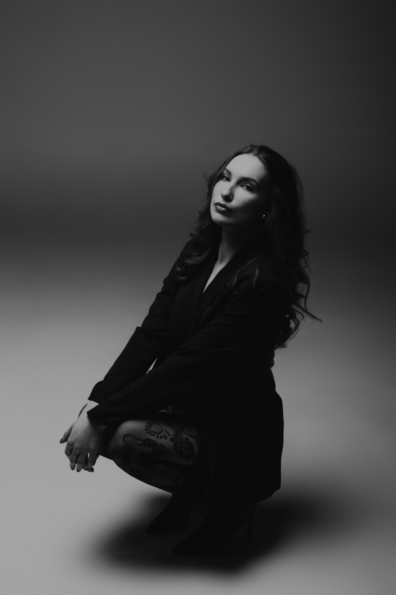 Czarno-białe portretowe ujęcie kobiety w ciemnym żakiecie i wzorzystych rajstopach, siedzącej na kucaka z rękami opartymi na kolanach, spoglądającej w górę.