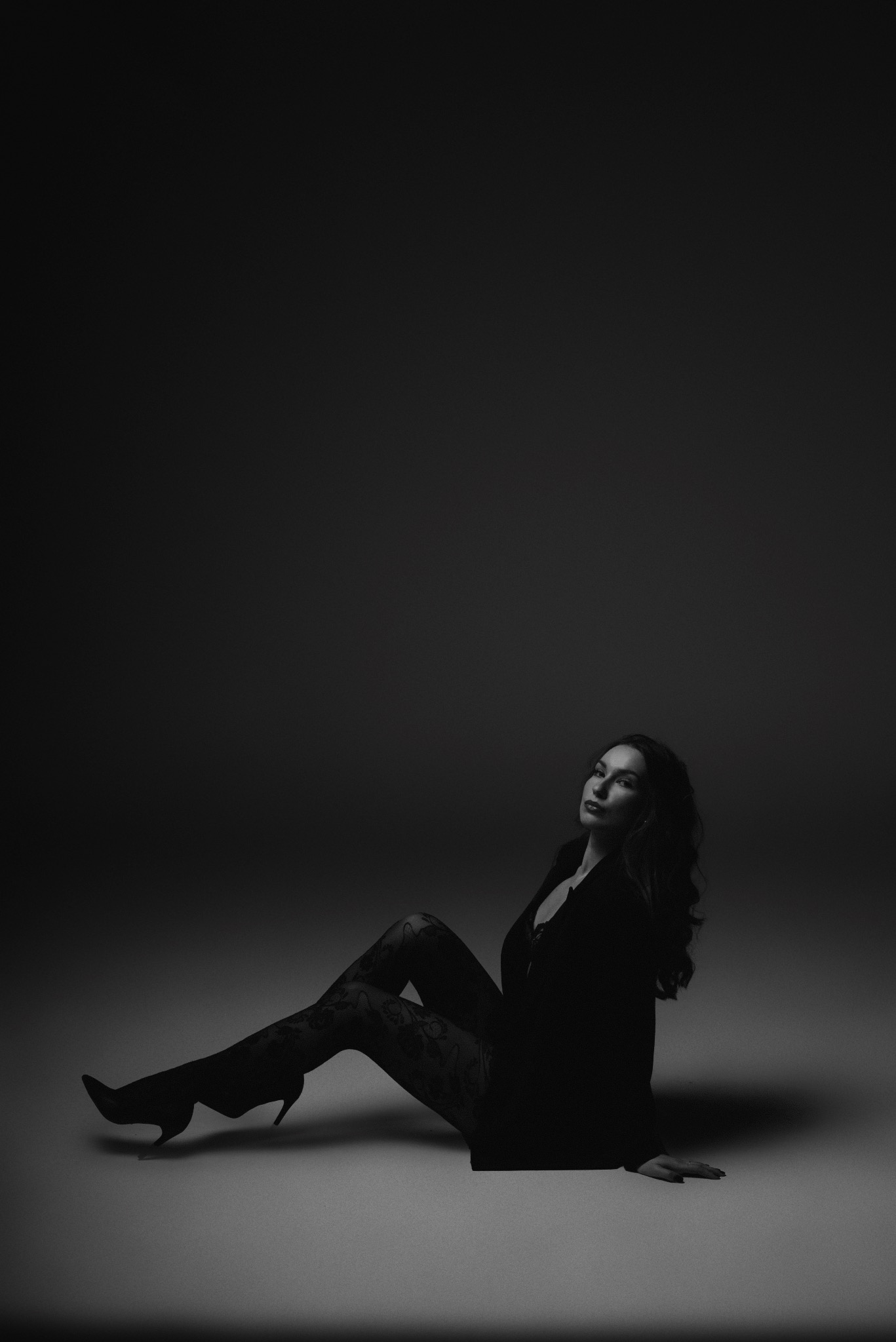 Kobieta w czarnej marynarce i wzorzystych rajstopach, siedząca na podłodze w studio, oświetlona od dołu, w monochromatycznej tonacji.